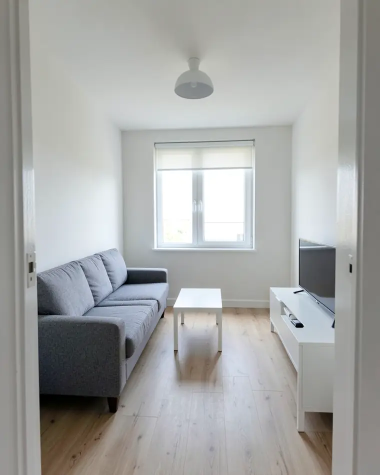 Schlichtes Wohnzimmer in einer Neubauwohnung: weiße Wände, ein graues Sofa, ein kleiner weißer Couchtisch, ein weißes TV-Board und ein Fenster mit Rollo