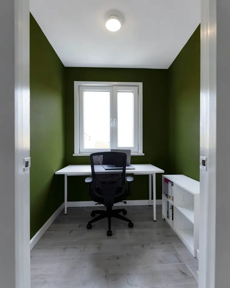 O mesmo home office com paredes em verde-oliva escuro e a mesma escrivaninha branca, cadeira preta e estante