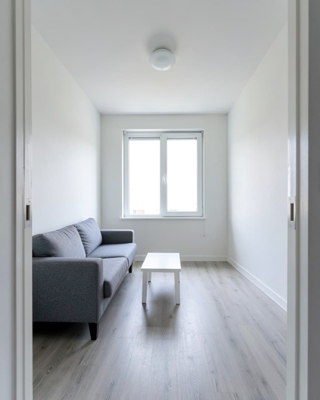 Before: a plain living room photo with white walls, a gray sofa, a white coffee table and a light floor, window straight ahead