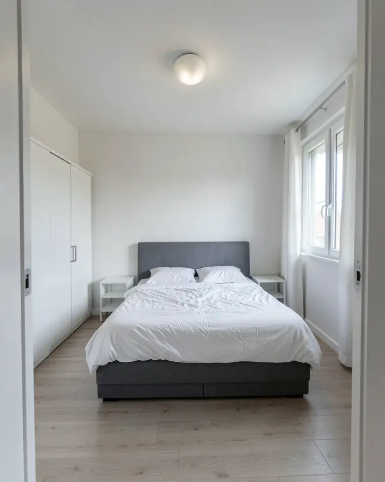 Camera da letto spoglia in un appartamento nuovo: pareti bianche, letto matrimoniale con testiera grigia e biancheria bianca, comodini bianchi, un armadio bianco e una finestra con tende bianche leggere