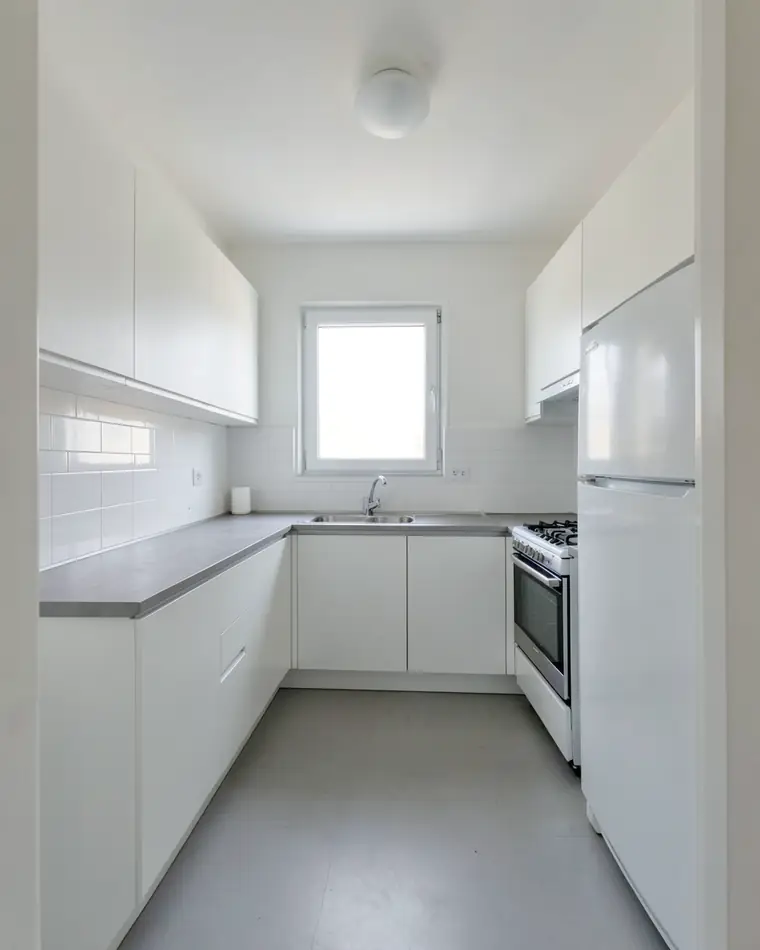 Plain white kitchen in a new apartment: white handleless cabinets, a gray countertop, white wall tiles and a window above the sink