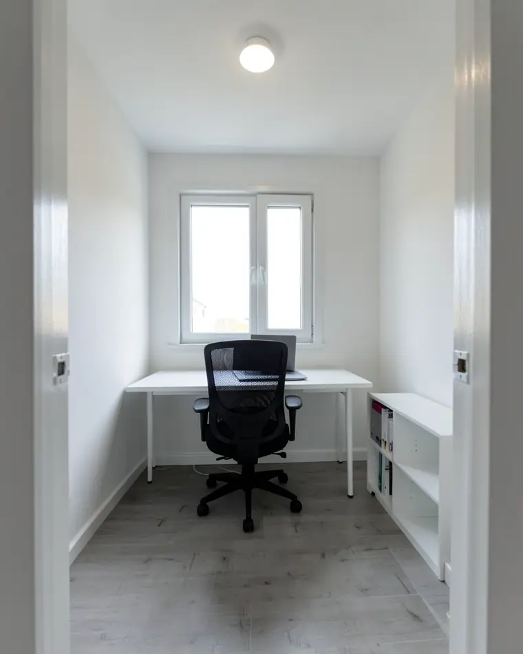Home office simples em um quarto pequeno de hóspedes: paredes brancas, escrivaninha branca embaixo da janela, cadeira de escritório preta e estante baixa branca